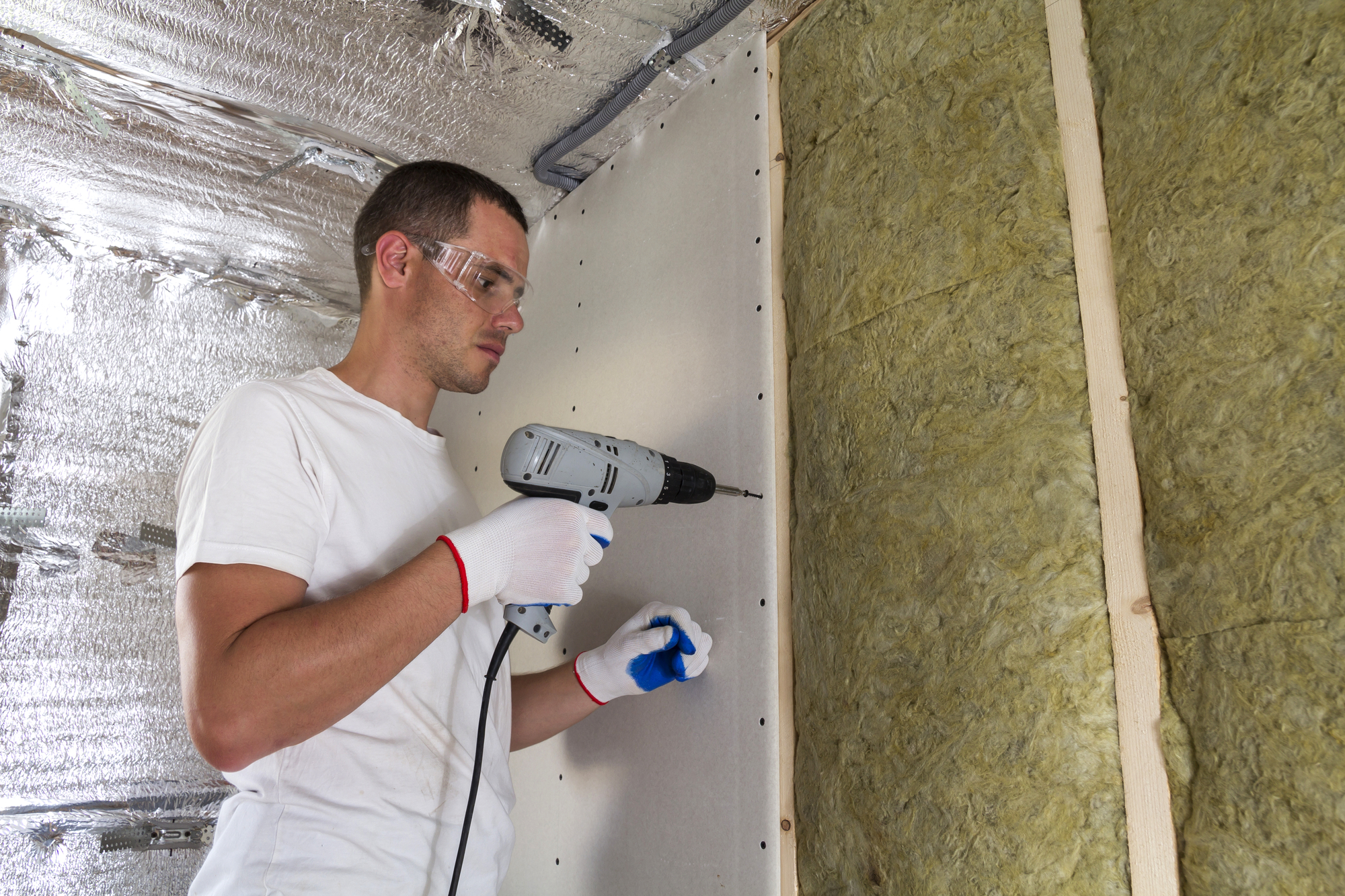 Worker in goggles with screwdriver working on insulation. Drywall on wall beams, insulating rock wool staff in wooden frame. Comfortable warm home, economy, construction and renovation concept.
